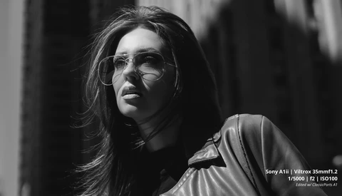 Person wearing clear-framed glasses and light-colored jacket, photographed in black and white with directional side lighting.