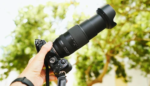 Photographer holding a telephoto lens mounted on a DSLR camera outdoors against a blurred green foliage background.