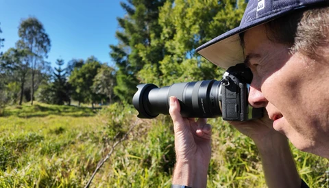 Photographer holding a telephoto lens camera to his eye while shooting outdoors in a forested area.