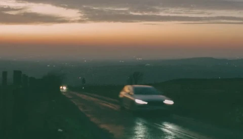 Two cars driving on a wet road at dusk with mountains silhouetted against a twilight sky.