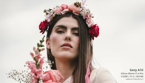 Woman with long dark hair wearing a elaborate floral crown of pink and red flowers with baby's breath, against a neutral background.