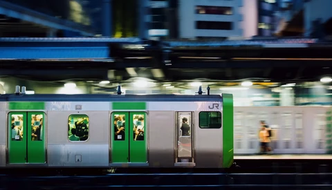 Motion-blurred train with green and silver livery moving through an urban station at dusk.