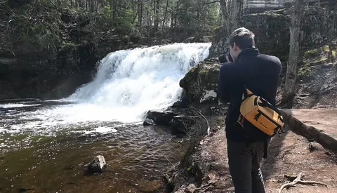 Photographer with camera and yellow bag documents a cascading waterfall in a forested stream setting.