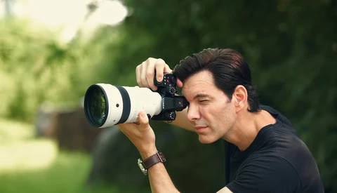 Photographer holding a telephoto lens camera to his eye, positioned in profile against a blurred green outdoor background.