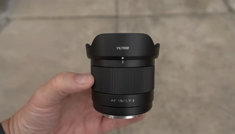 Hand holding a Viltrox AF 15/1.7 E lens with hood attached against a concrete background.