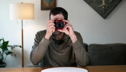 Photographer holding a red digital camera up to his face while seated at a wooden table.