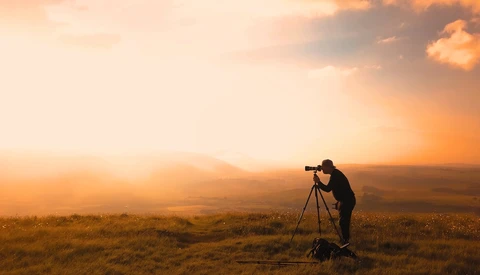 Photographer silhouetted against golden sunset, aiming telephoto lens on tripod across expansive landscape.