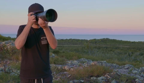 Photographer holding a telephoto lens to his eye while standing on rocky terrain at dusk.