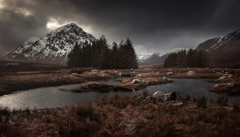 Snow-capped mountain peak rising above forested valley with partially frozen stream in foreground under dramatic cloudy sky.