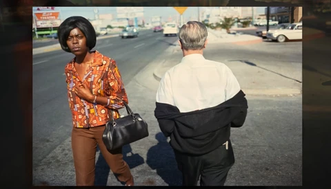Two people standing on a sunny street with parked cars visible in the background.