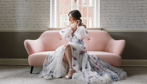 Woman in white blouse and floral skirt sitting on pink sofa near window, touching her face thoughtfully.