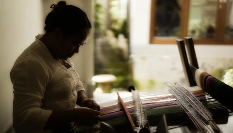 Person working on a weaving loom with colorful yarn threads in soft, directional window light.