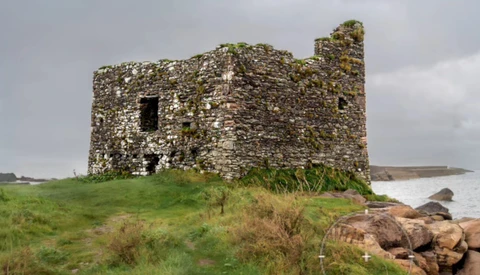 Stone ruins of a multi-story structure with moss-covered walls and empty window openings on a grassy hilltop overlooking water.