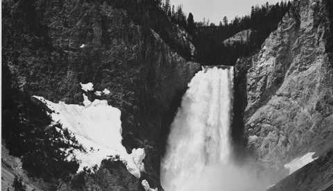 Powerful waterfall cascading down a rocky canyon with forested cliffs and patches of snow.