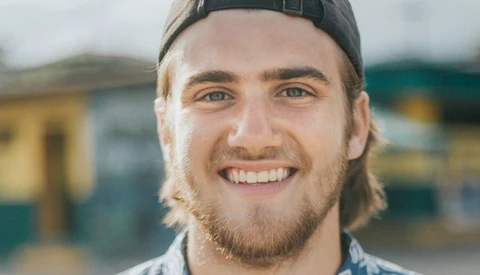 Smiling man with shoulder-length hair and beard wearing a black cap and light colored shirt outdoors.