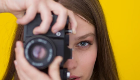 Young woman holding a vintage film camera up to her eye against a bright yellow background.