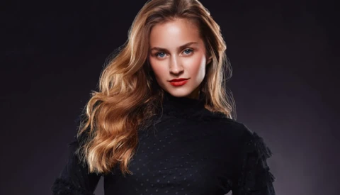 Woman with golden-brown wavy hair wearing a black turtleneck against a dark background.