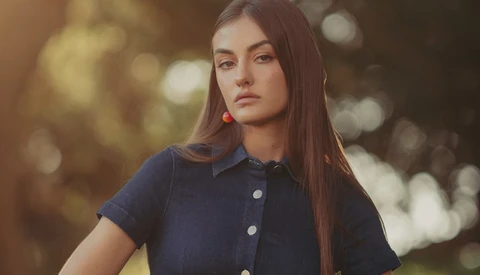 Young woman with long dark hair wearing a navy button-up shirt and red earrings, photographed outdoors with soft golden bokeh background.