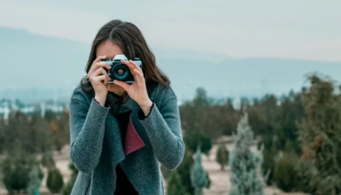 Woman holding a camera up to her face while standing outdoors with forested mountains in the background.