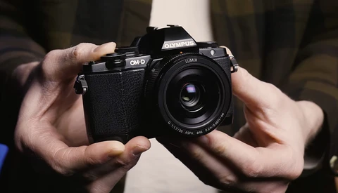 Hands holding an Olympus OM-D mirrorless camera with a Lumix prime lens attached.