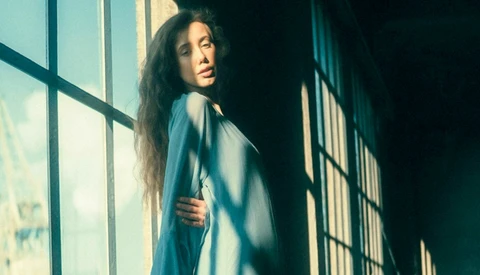 Woman with long dark hair looking over her shoulder by a sunlit window with grid panes.