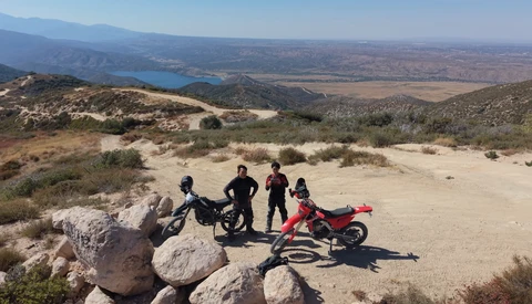 Four people with two motorcycles standing on rocky terrain overlooking a vast landscape with a blue lake in the distance.