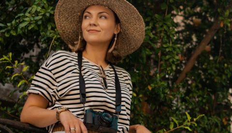 Woman in striped shirt wearing straw hat and holding camera with neck strap in front of green foliage.