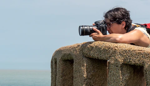 Photographer shooting with a telephoto lens while perched on a concrete structure against a clear sky.
