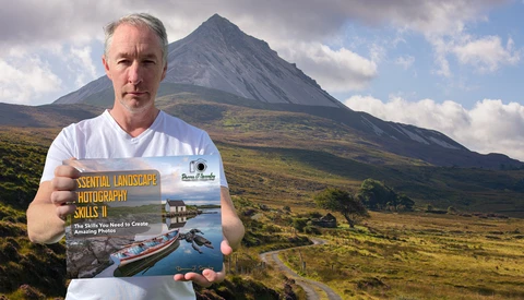 Photographer holding a landscape photography instructional book with a dramatic conical mountain and moorland in the background.