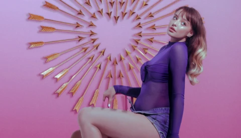 Woman in purple outfit posing against pink background with gold arrow wall decoration.