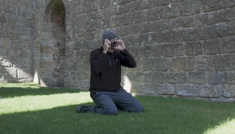 Photographer kneeling on grass against stone fortress wall, holding camera to eye to compose a shot.
