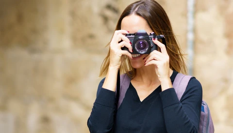 Woman holding a compact mirrorless camera to her face while standing against a textured stone wall.