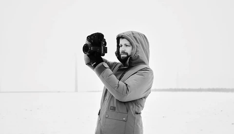 Bearded man in hooded jacket holding vintage film camera to his face against minimal background.