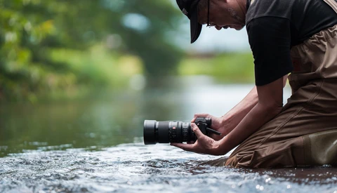 Photographer crouching in shallow water while holding a camera with telephoto lens.