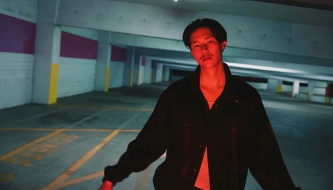 Man in black jacket standing in empty parking garage with red and yellow lighting.
