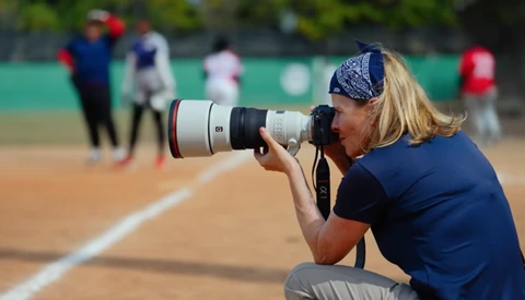 Sports photographer crouching on clay court with telephoto lens during outdoor baseball game.