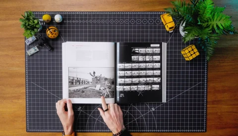 Photographer's hands holding an open book displaying black and white photographs on a grid-patterned cutting mat.