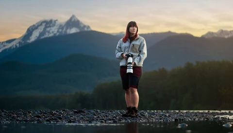 Person standing in shallow water holding a camera, with a snow-capped mountain peak in the background at golden hour.