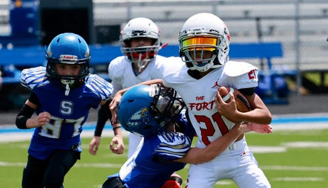 Youth football player in white uniform carrying ball while being tackled by defender in blue uniform.