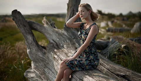 Woman in floral dress posing on weathered driftwood in an open field.