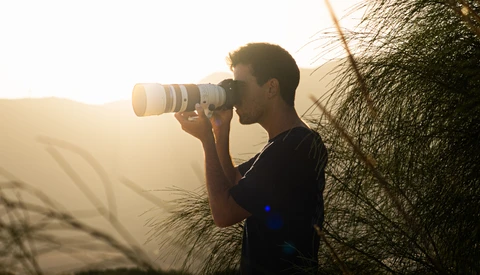 Photographer with telephoto lens shooting into golden sunset light near dried grass and tree branches.