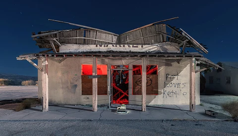 Abandoned roadside diner with boarded red doors and deteriorating wooden roof against clear blue sky.