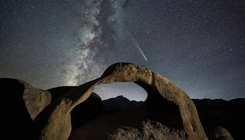 Milky Way galaxy arching over a natural rock arch formation under a starry night sky with a visible meteor.