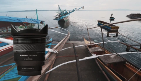 Schneider lens displayed in foreground with fishing boats anchored in calm coastal waters behind it.