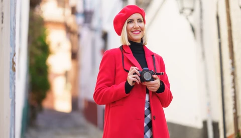 Woman in bright red coat and beret holding vintage camera on narrow European street.