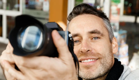 Male photographer holding a camera with a large telephoto lens, smiling at the camera.