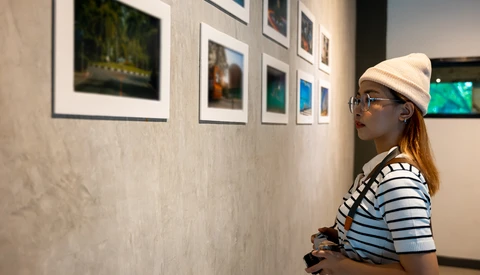 Woman in striped shirt and cream cap viewing framed photographs on gallery wall while holding camera.