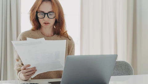 Woman with reddish hair and glasses reviewing documents while working at laptop at desk.