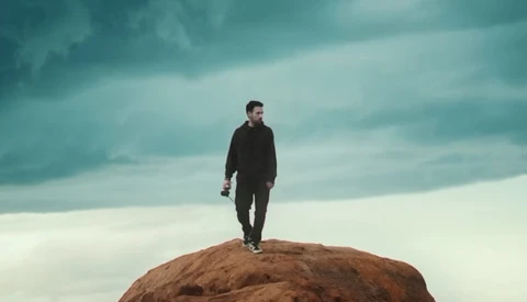 Man in dark clothing standing on top of a large rocky boulder against a dramatic cloudy sky.