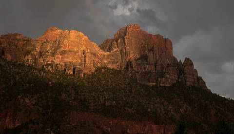 Red rock formations illuminated by golden sunlight beneath a dramatic stormy sky.